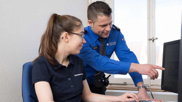 Zwei Personen, die für das BAZG arbeiten der Mann im blauen Hemd zeigt der Frau im schwarzen T-Shirt etwas auf dem Computer